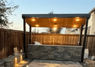 A modern stone outdoor bar under a black pavilion with a warm wood ceiling, featuring recessed LED lighting and illuminated stone walkway pavers.