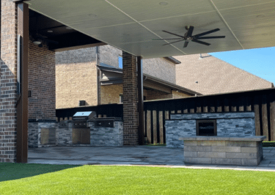 Large modern covered patio featuring an outdoor kitchen with grey stone veneer, a built-in grill, a wine cooler, a square stone fire pit, and a lush artificial turf lawn.