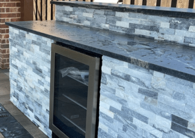 Close-up of a custom outdoor kitchen island featuring grey and white stacked stone veneer, a black granite countertop, and a built-in stainless steel beverage cooler.