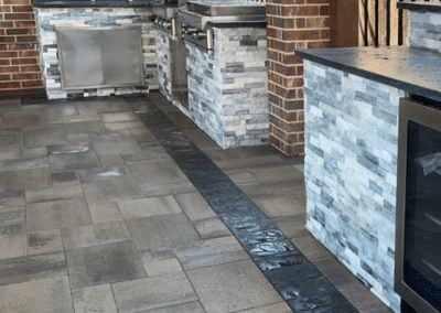 A professional outdoor kitchen featuring grey stacked stone cabinetry, a built-in stainless steel gas grill with a side burner, a sink station, and a beverage cooler on a paver patio.
