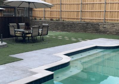 Modern backyard with a geometric swimming pool, a light grey stone patio, and a dining set with a large umbrella on a vibrant artificial turf lawn.