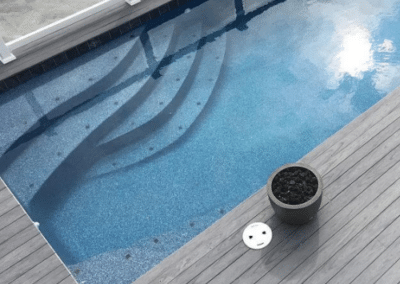 Overhead view of a rectangular swimming pool featuring blue speckled plaster, curved integrated stairs, and a grey wood-grain composite deck with a glass safety railing.