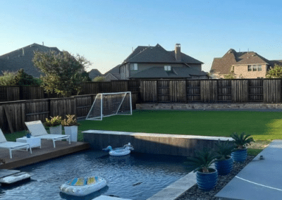 Modern backyard with a large swimming pool, an elevated white spa, a wooden sun deck with loungers, and a professional artificial turf lawn featuring a soccer goal.