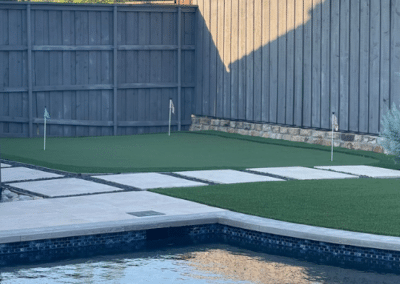 Modern backyard featuring a professional-grade artificial turf putting green with three flags next to a luxury swimming pool and a geometric stone paver walkway.
