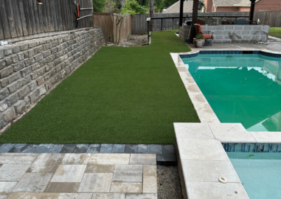 A modern backyard featuring a luxury swimming pool, a custom wood-framed gabled pavilion, and a vibrant artificial turf lawn bordered by a stone retaining wall and multi-tonal pavers.