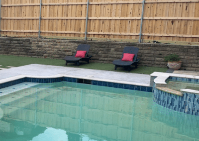 Modern geometric swimming pool featuring a raised blue-tiled spa, sun loungers with red accent pillows on an artificial turf lawn, and a tiered stone retaining wall.